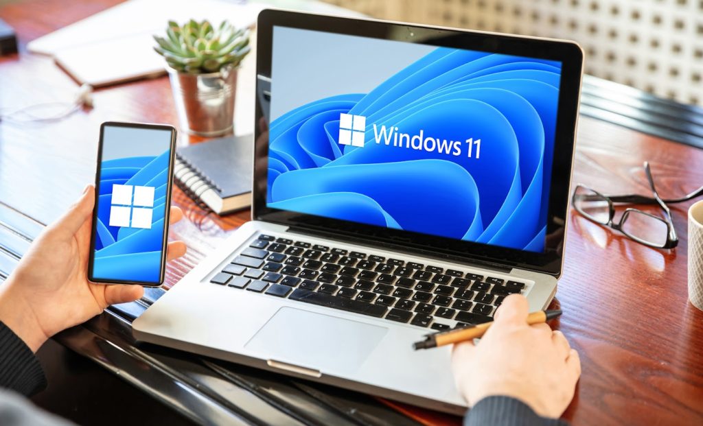 An image of a man's hands with a laptop displaying Windows 11 operating system on the screen and a smartphone with a business office desk background.