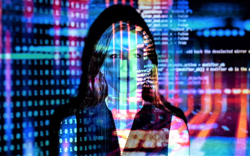 Image of a woman with bright fluorescent lights shining in her face, the lights are projecting code and analytics.