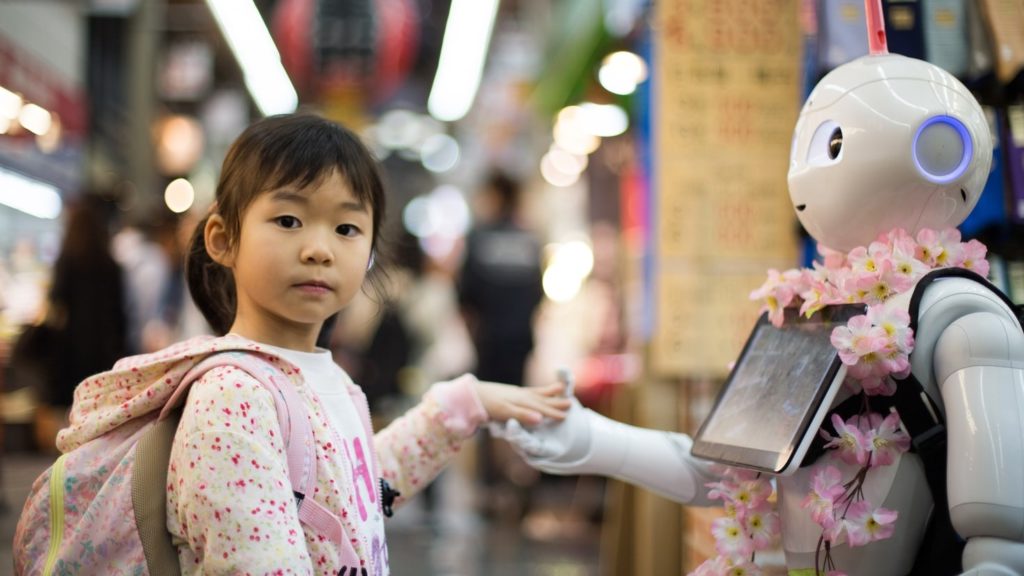 Image of a little girl interacting with a robot. The girl is holding the robot's hand.
