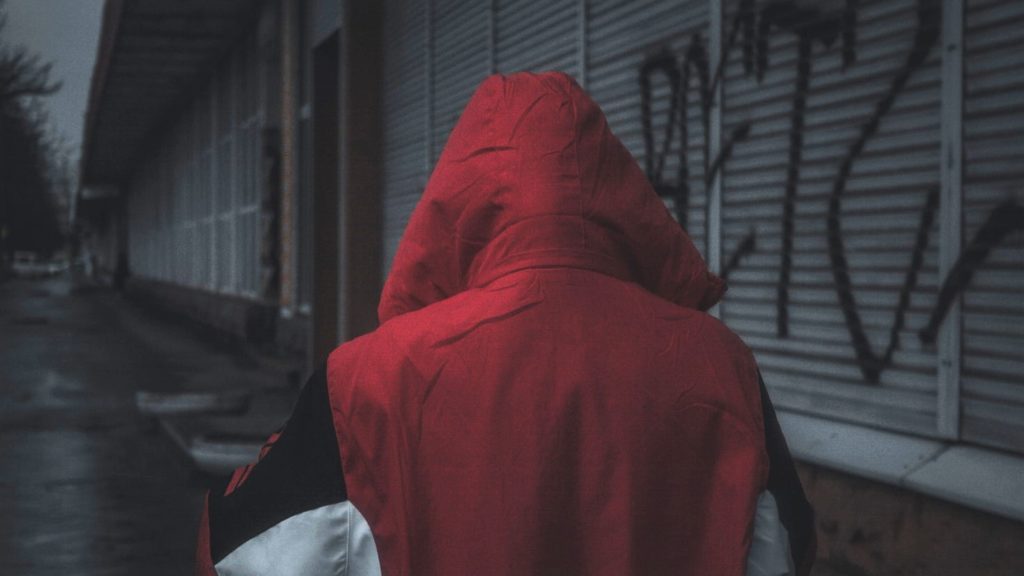 Image of a hooded person in a red coat walking in an alley.