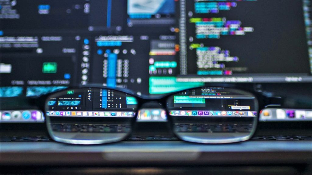 Image of glasses on a desk. Through the lenses, we see several monitors and screens.