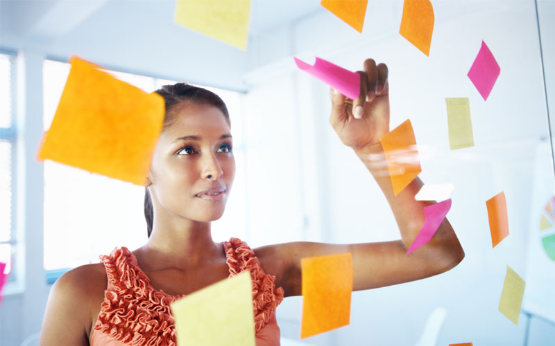 A lady facing a transparent glass pane bearing multiple colored post its. The lady is peeling off one post-it.