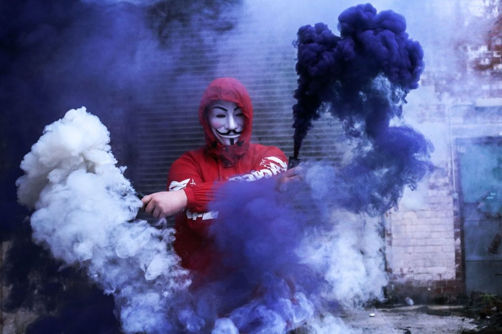 Image of a guy in a red hoodie and Guy Faux mask  setting off smoke bombs.