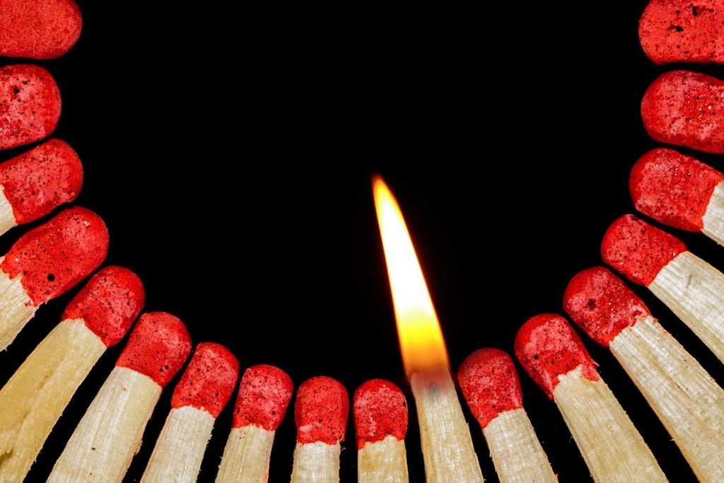 An image of a circular row of matches against a black background. One match has been lit.