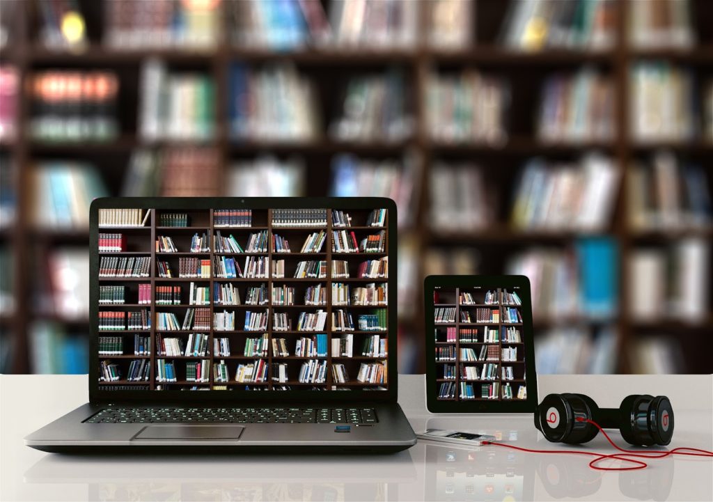 Image of a laptop and a tablet on a table in front of a bookshelf filled with books. The laptop and tablet display images of a bookshelf filled with books on their screens.