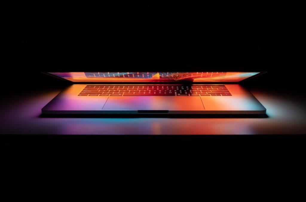 Image of a laptop slightly open, emitting colorful lights in a dark space.