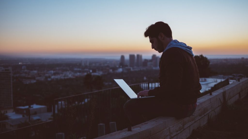 Image of a man typing on a laptop while sitting on a ledge overlooking the city.