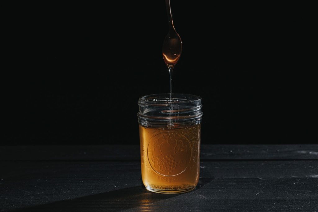 mage of a jar of honey on a table with a spoon above it, dripping honey.