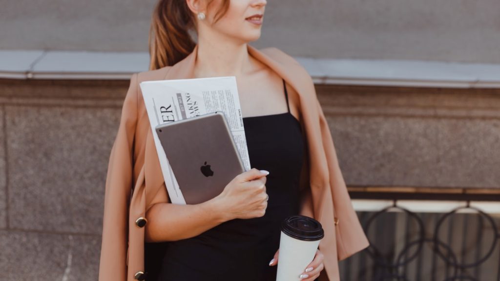 Image of woman in a tan coat holding a tablet with newspapers in one hand and a coffee in the other hand.