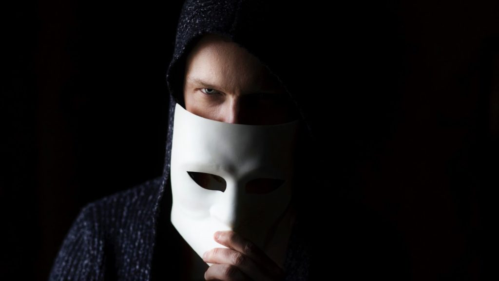 Image of a man wearing a hoodie is hiding half of his face behind a white mask.