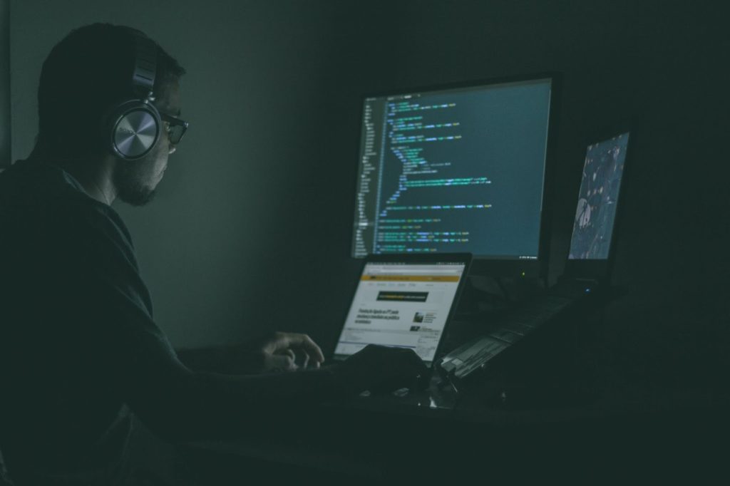 An image of a man wearing headphones and glasses is working on three different-sized monitors in a dark room