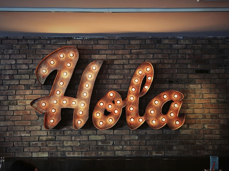 Image of a lit Hola sign on a brick wall.
