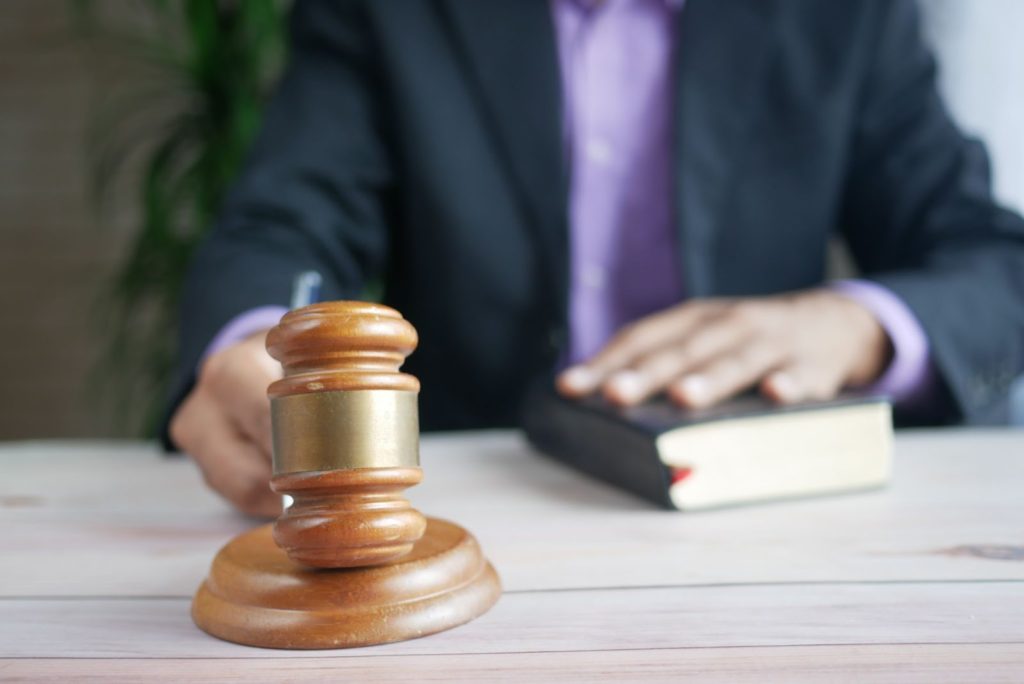 The image shows a legal official dressed in a suit holding a gavel on a flat, wooden surface. 