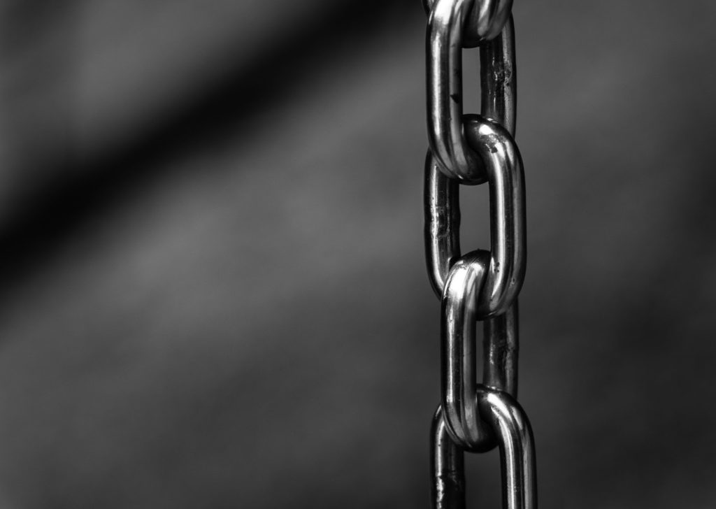 The image shows a shiny silver or steel chain against a grey background, right from center in the frame.