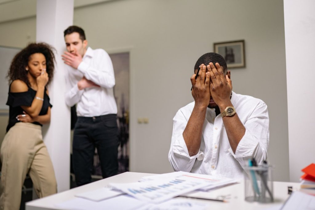 The image shows a man in a white dress shirt sitting at a desk covering his face, while two of his colleagues stand in the background