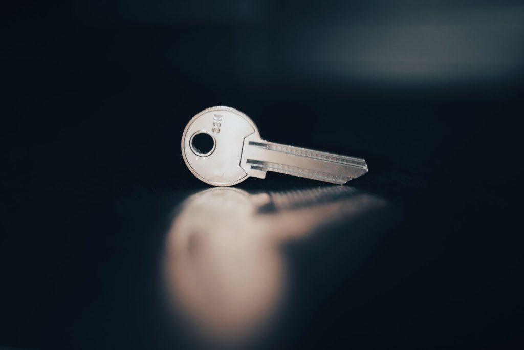 A silver key against a black background. Slight blurry reflective effect on the bottom aspect of the picture. 