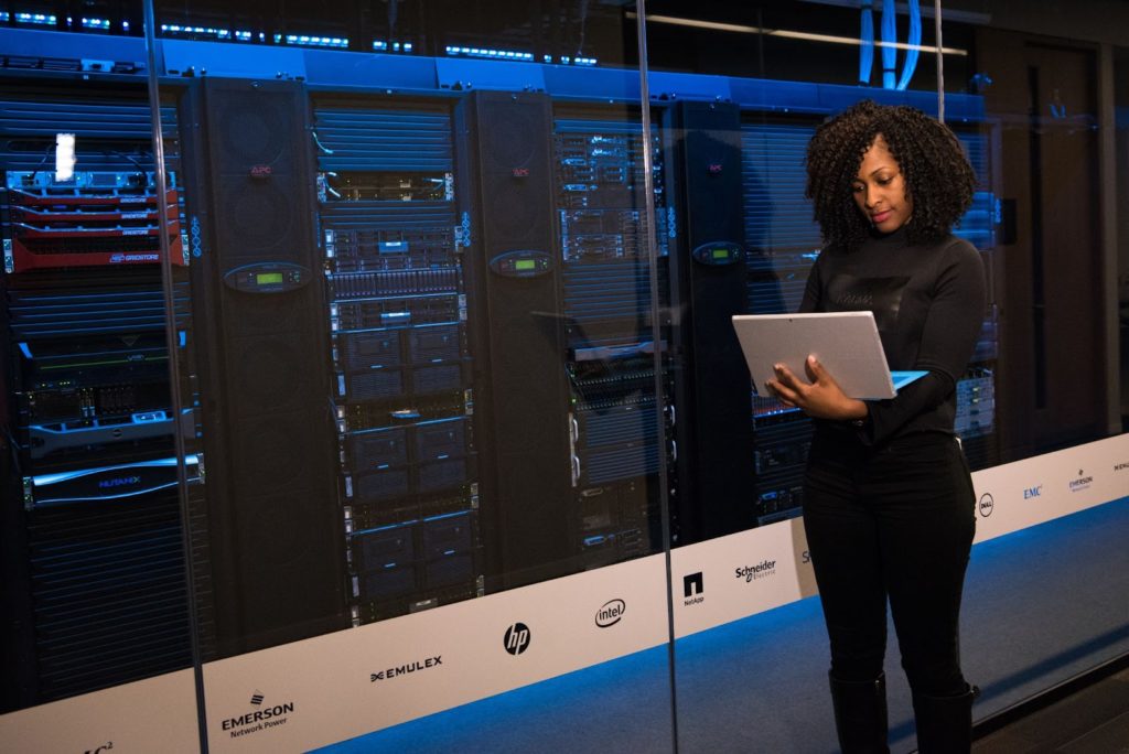 The image shows a woman in black, holding a laptop,
standing against three large servers enclosed behind glass with blue lighting.