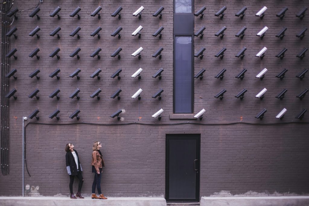 The image shows lots of black and white CCTV cameras against a brick wall with two women looking up toward them.